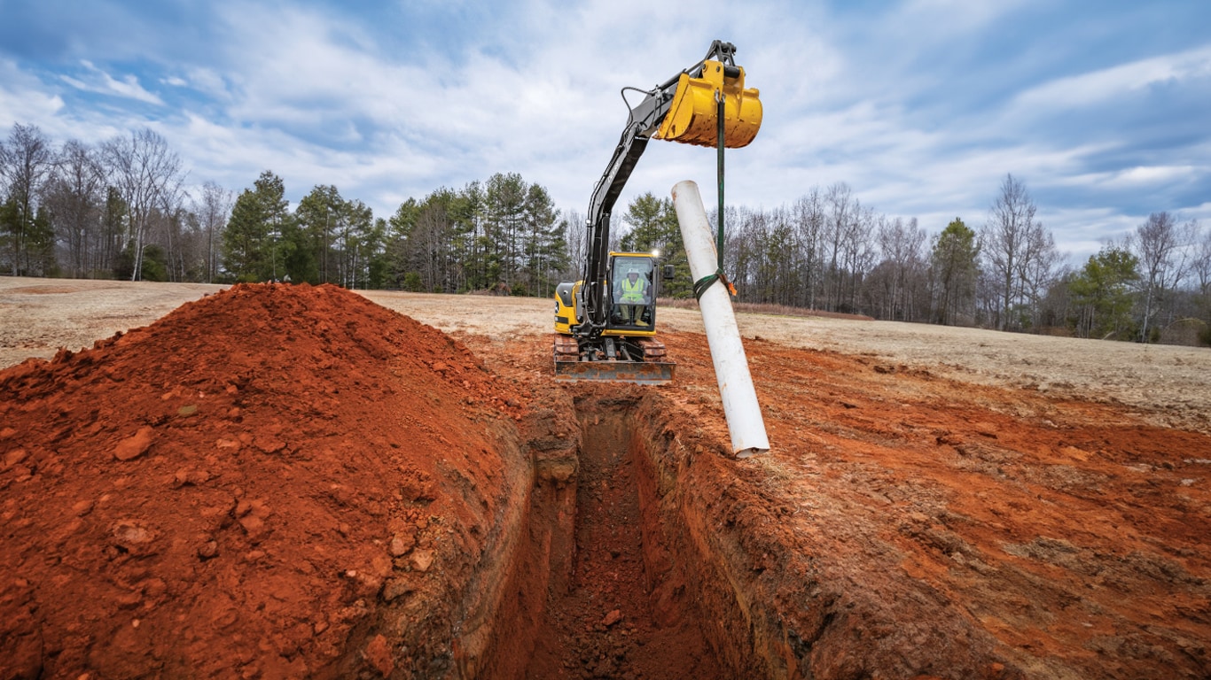 A John Deere Excavator lowering a white pipe into a trench on a worksite.